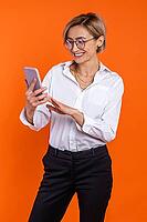 Joyful beautiful woman wearing white official style shirt using mobile phone isolated over orange background [IBR123569293]