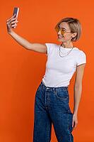 Satisfied woman in white t shirt and jeans making selfie broadcasting livestream isolated over orange background [IBR123569298]