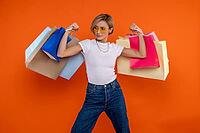 Funny strong woman in white t shirt and jeans raised arms with shopping bags isolated over orange background [IBR123569299]
