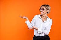 Delighted woman wearing white official style shirt presenting copy space on palm isolated over orange background [IBR123569302]