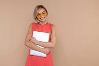 Confident happy beautiful woman in dress and sunglasses holding laptop empty space isolated over beige background [IBR123569303]