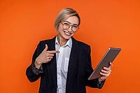 Happy woman wearing black official style suit pointing at tablet isolated over orange background [IBR123569307]