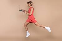 Jumping woman in dress and sunglasses using laptop working online isolated over beige background [IBR123569311]