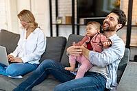 Father carrying sleeping daughter while looking at woman using laptop on sofa [IBR123569319]