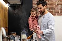 Father cooking meal holding toddler daughter in kitchenn preparing soup [IBR123569322]