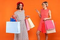 Satisfied women friends with shopping bags enjoying new purchases isolated over orange background [IBR123569324]
