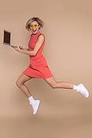 Jumping woman in dress and sunglasses using laptop working online isolated over beige background [IBR123569327]