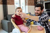 Bearded father feeding little baby daughter sitting in highchair with puree or porridge by spoon at home [IBR123569331]