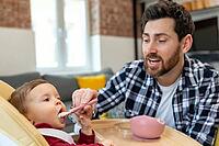 Brunette father teaching his little baby to eat porridge from spoon infant daughter sitting in highchair [IBR123569333]