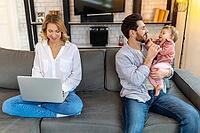 Father carrying infant daughter while mother using laptop on sofa remote work husband helping his wife with baby child [IBR123569334]