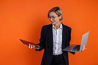 Joyful woman wearing black official style suit choosing laptop or tablet isolated over orange background [IBR123569337]