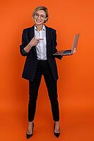 Confident smiling woman wearing black official style suit pointing at laptop in her hands isolated over orange background [IBR123569338]