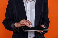 Unrecognizable woman wearing black official style suit using tablet isolated over orange background [IBR123569341]