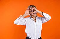 Romantic woman wearing white official style shirt looking through heart shape with hands isolated over orange background [IBR123569342]