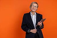 Confident satisfied woman wearing black official style suit standing with tablet looking at camera isolated over orange background [IBR123569343]