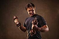 Young man hardly doing exercises with dumbbells on stone wall background. Stress and effort emotions [IBR123569357]