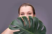 Smiling caucasian adult female peeking from monstera leaf against gray background [IBR123629728]