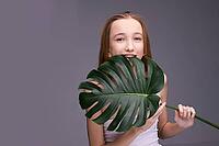 Young caucasian female teen with long hair holding large monstera leaf against gray background and smiling [IBR123629729]