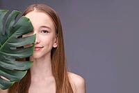 Young caucasian female with long hair holding leaf in minimalist portrait on gray background [IBR123629730]