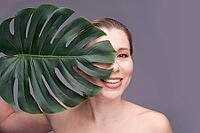 Caucasian female adult smiling behind monstera leaf on gray background [IBR123629731]