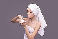 Young asian female applying skincare cream with towel on head against gray background [IBR123629732]