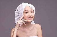 Young caucasian female skincare routine smiling with towel and cream on face against gray background [IBR123629734]
