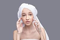 Young caucasian female with under eye patches and towel on head, gray background, skincare routine [IBR123629736]
