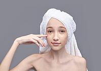 Young asian female applying under eye patch with towel on head for skincare routine on gray background [IBR123629744]