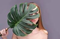 Young caucasian female partially covered by large green leaf against gray background [IBR123629745]