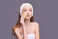 Young caucasian female with spa headband cleansing skin using facial sponge against gray background [IBR123629746]