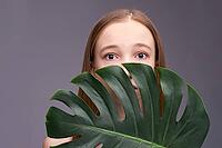 Young caucasian female with monstera leaf against gray background, expressive eyes, natural beauty [IBR123629747]