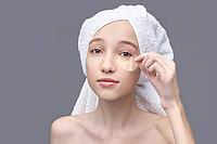 Young caucasian female applying eye patch with towel wrapped hair in skincare routine on grey background [IBR123629749]