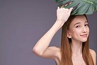 Young caucasian female holding leaf with playful smile in minimalist setting [IBR123629758]