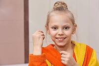 Smiling caucasian female child in bright bathrobe practicing dental hygiene with floss in bathroom setting [IBR123629759]