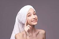Young caucasian female applying skincare cream with towel wrap on head smiling [IBR123629761]