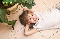 Smiling caucasian young girl relaxing on tiled floor next to potted green plants indoors [IBR123629767]