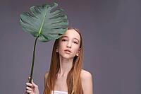 Young caucasian teen female holding large leaf against gray background with long straight hair and natural expression [IBR123629770]