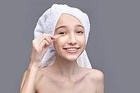 Young caucasian female applying under eye patch with towel on head against gray background [IBR123629771]