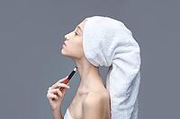 Young caucasian female applying makeup with brush on neck wrapped in towel against gray background [IBR123629773]
