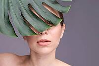 Caucasian female with monstera leaf partially covering face, close up portrait on gray background [IBR123629776]