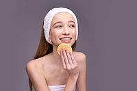 Smiling caucasian female teen using facial sponge for skin care routine with headband on grey background [IBR123629777]