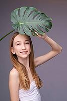 Young caucasian female holding monstera leaf in studio on gray background with long blonde hair and white top [IBR123629778]