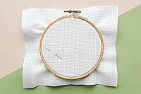 Wooden embroidery hoop with empty white canvas cloth and needle on pastel background with light pink and light green colors, ready for cross-stitching [IBR123638370]