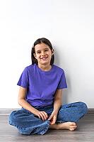Smiling white brunette girl aged 7-8 in purple t-shirt and jeans sitting barefoot on wooden floor against white wall [IBR123638371]