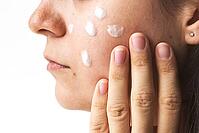 Close-up of young woman applying skincare cream to her cheek with fingers, visible texture of skin and cream on white background, concept beauty, skincare, self-care [IBR123638376]