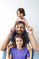 Father with two daughters posing together, smiling having fun. younger girl sits on his shoulders, while older one holds his hands. Happy moment in bright interior, concept of fatherhood, vertical [IBR123638377]
