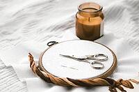 Embroidery hoop with white fabric, vintage scissors, and a needle lying on top, accompanied by a burning candle in a cozy setting. Aesthetic composition of handmade craft and relaxation [IBR123638379]