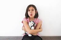 Child girl 7-8 years in pink t-shirt sits against white wall, hugging plush toy and sticking out her tongue with stubborn expression. She appears playful and defiant. Concept of childhood emotions [IBR123638383]