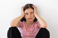 A young girl 7-8 year in a pink shirt sits against a white wall, rolling her eyes and holding her head in frustration, symbolizing boredom, annoyance, or stress [IBR123638387]