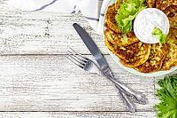 Fritters from zucchini with sour cream sauce and greens. Vegetable pancakes of courgettes with green salad on white wooden background. Top view with copy space [IBR123639579]